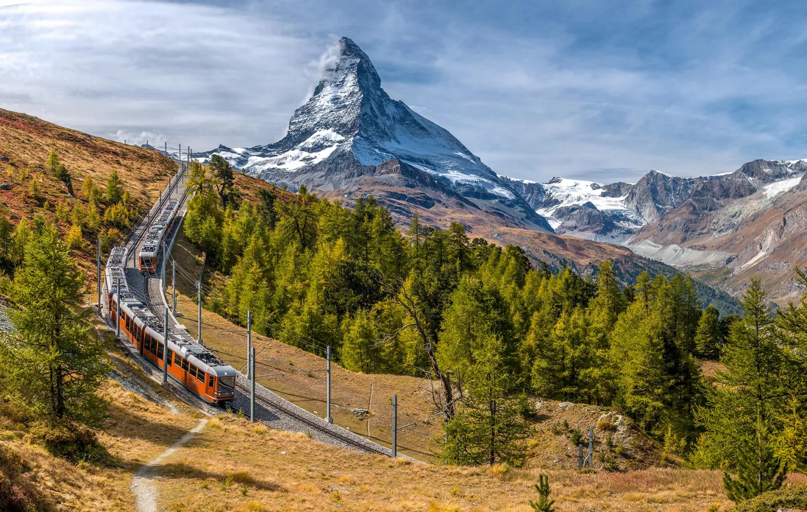 A red Gornergrat cogwheel train climbing through autumn larch below the Matterhorn