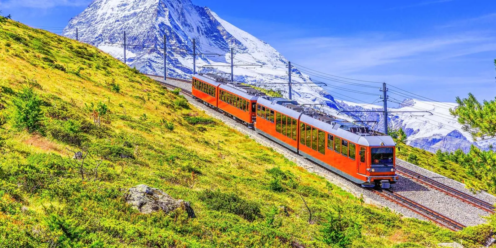A red Gornergrat train on a green alpine slope beneath snow-covered peaks