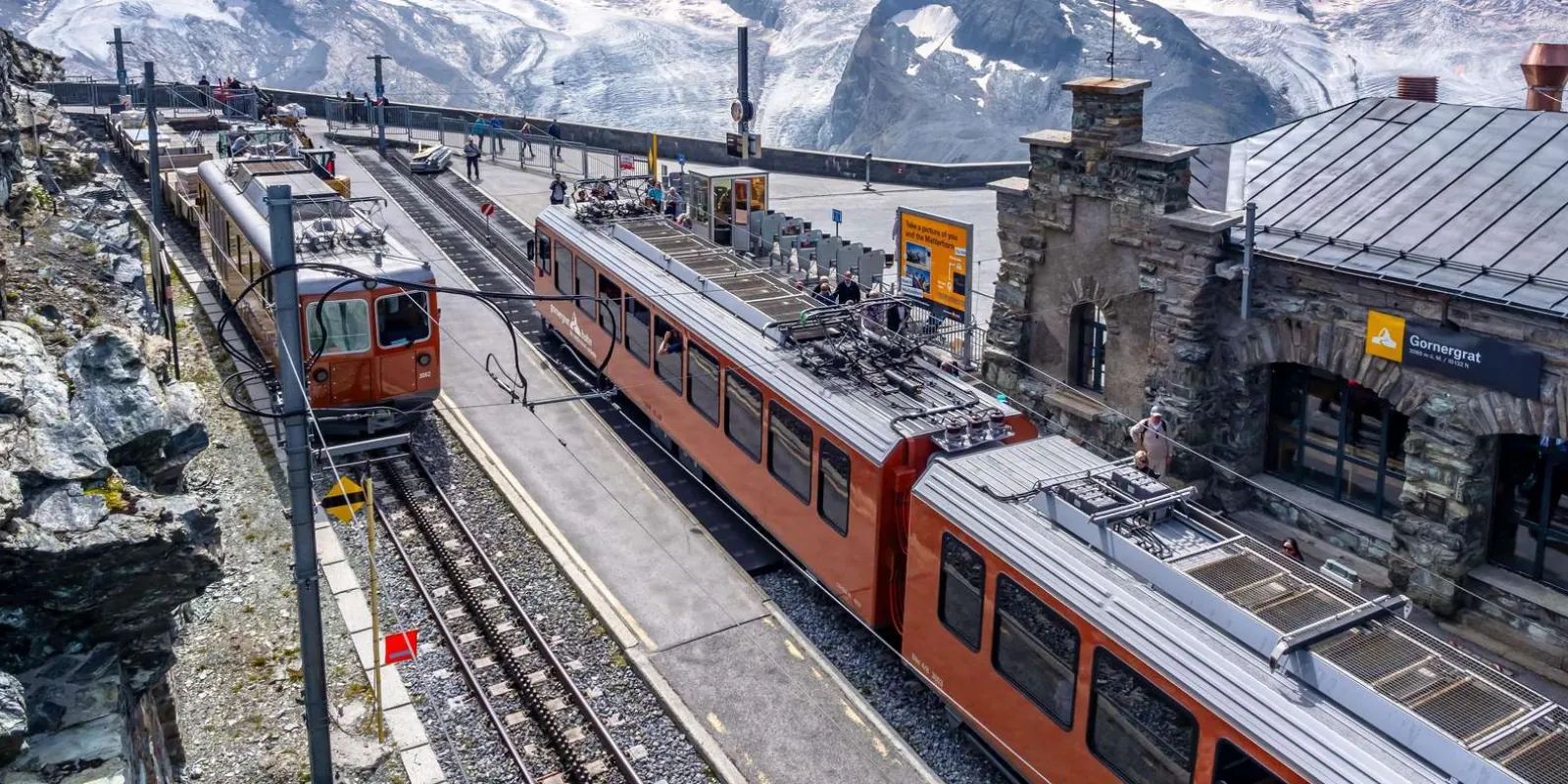 Two red Gornergrat cogwheel trains at the summit station platform beside the yellow Gornergrat 3,089 m altitude sign
